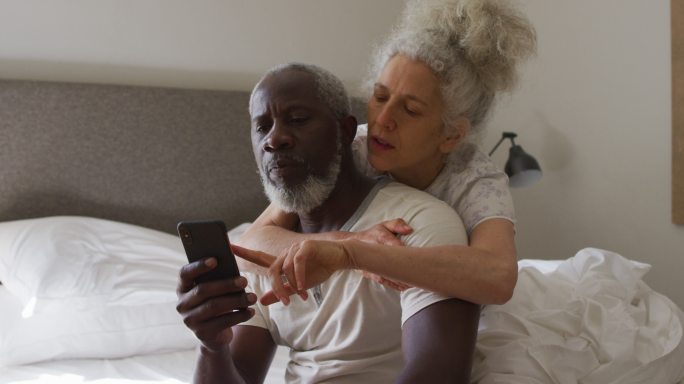 混合种族的老年夫妇坐在家里的床上使用智能手机