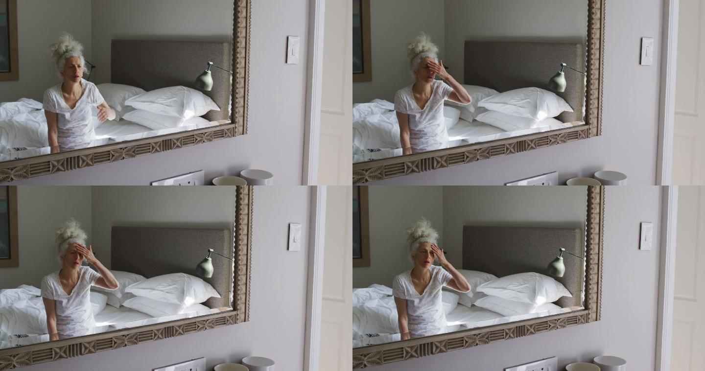 一位年长的白人妇女坐在家里的床上看着镜子摸着自己的脸