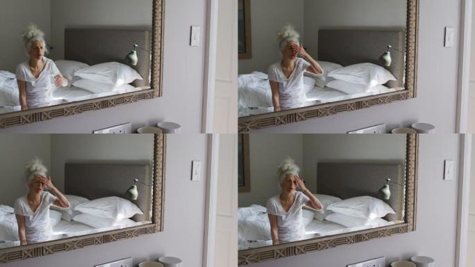 一位年长的白人妇女坐在家里的床上看着镜子摸着自己的脸