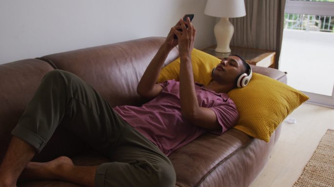 混血男子戴着耳机躺在沙发上使用智能手机