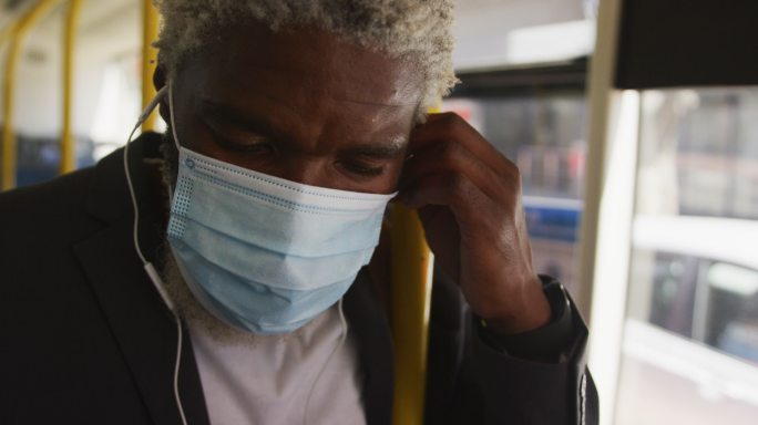 非裔美国老人戴着口罩，戴着耳机站在公共汽车上