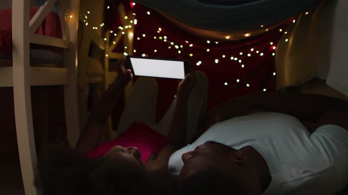 一个非裔美国人的女儿和她的父亲躺在有仙女灯的毯子帐篷里看平板电脑