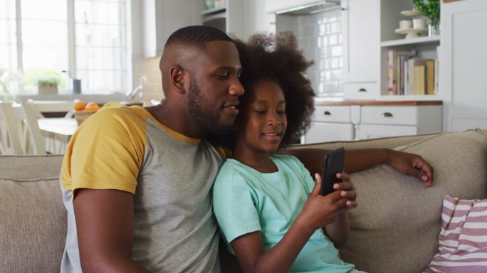 非裔美国人的女儿和她的父亲一起坐在沙发上使用智能手机