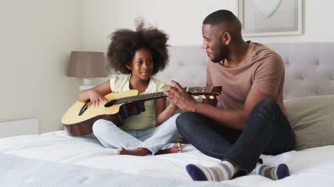 一位非裔美国父亲和他的女儿坐在床上一起弹吉他