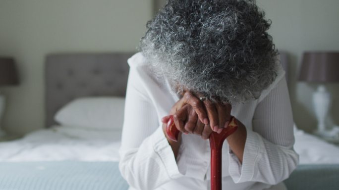 一位年长的非裔美国妇女拄着拐杖坐在家里的床上