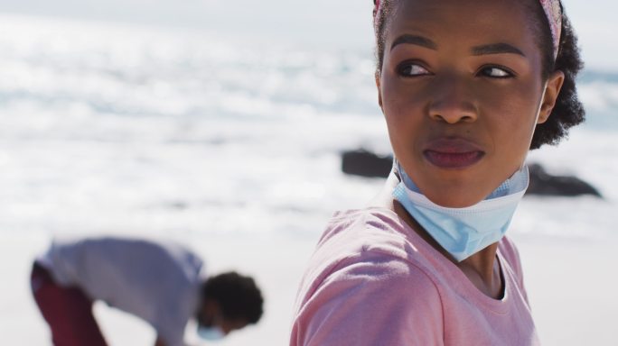 一位戴着面罩的非裔美国妇女在海滩上捡垃圾时休息