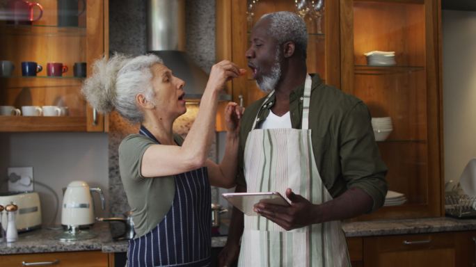 在家里的厨房里，穿着围裙给丈夫喂切碎的蔬菜的白人老年妇女