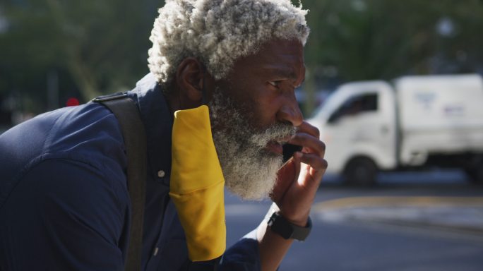 非洲裔美国老人戴着口罩在路上用智能手机讲话的特写
