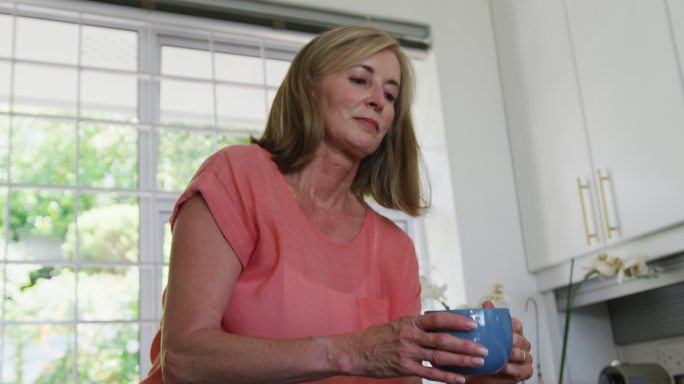 一个白人高级妇女站在家里的厨房里喝着咖啡