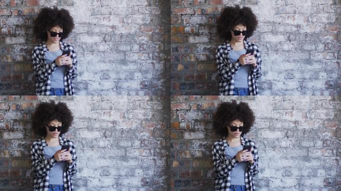 非裔美国妇女戴着太阳镜，靠砖墙站着用智能手机