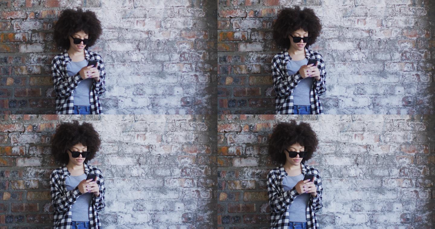 非裔美国妇女戴着太阳镜，靠砖墙站着用智能手机