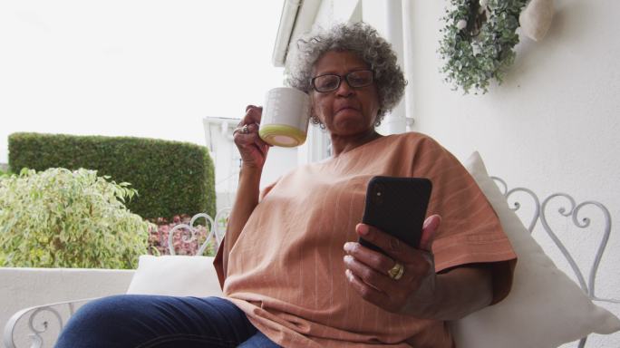高级非洲裔美国妇女一边用智能手机一边喝咖啡，坐在侯的门廊上