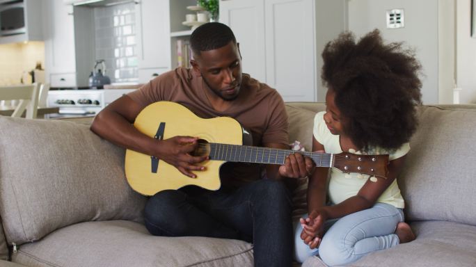 一位非裔美国父亲和他的女儿坐在沙发上弹吉他