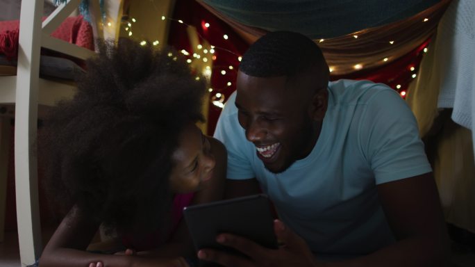 非洲裔美国人的女儿和她的父亲在有仙女灯的毯子帐篷里看平板电脑