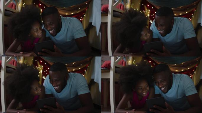 非洲裔美国人的女儿和她的父亲在有仙女灯的毯子帐篷里看平板电脑