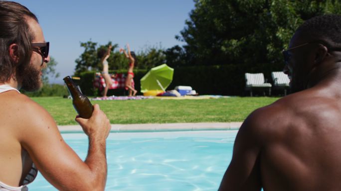 两个不同肤色的男性朋友坐在泳池边敬酒和喝啤酒