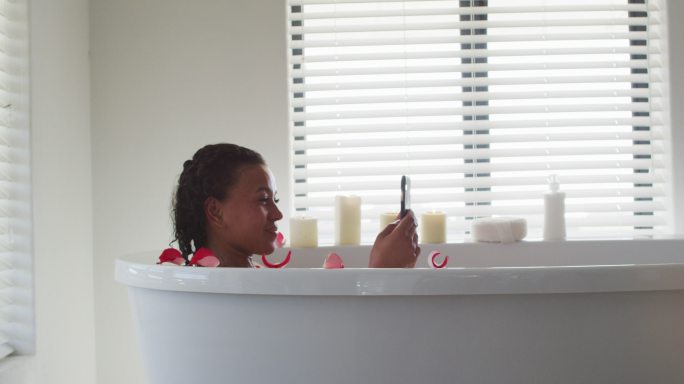 非裔美国女性在家里浴室的浴缸里使用智能手机