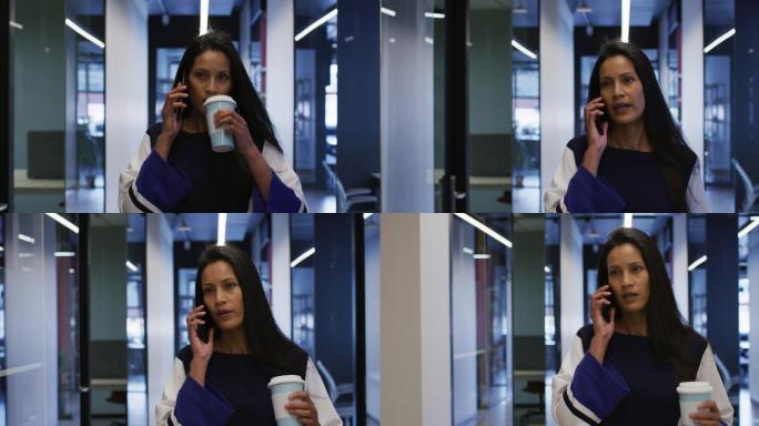 混血女商人拿着咖啡在办公室走廊边走边用智能手机聊天