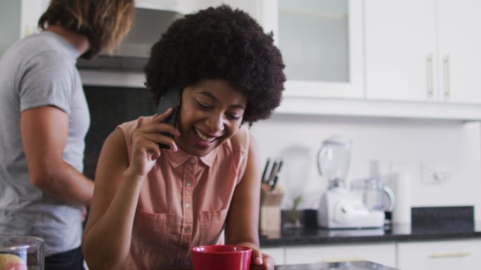 一位非裔美国女性拿着咖啡杯在家厨房用智能手机聊天