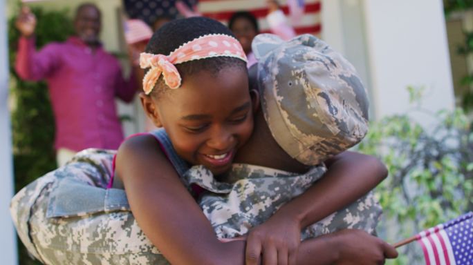 非裔美国士兵父亲拥抱微笑的女儿，身后是家人和美国国旗