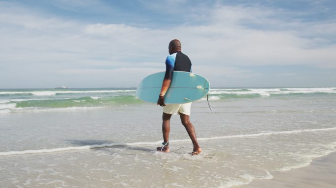 非裔美国老人在沙滩上走着，手里拿着冲浪板，往海里跑