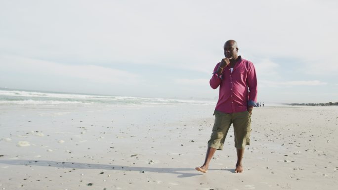 非裔美国老人在沙滩上走着，揉着下巴，看着大海