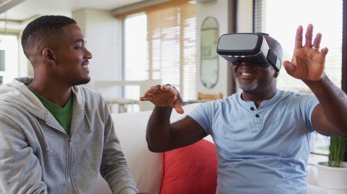 一位非裔美国老人坐在沙发上，戴着虚拟现实头盔，他十几岁的儿子面带微笑