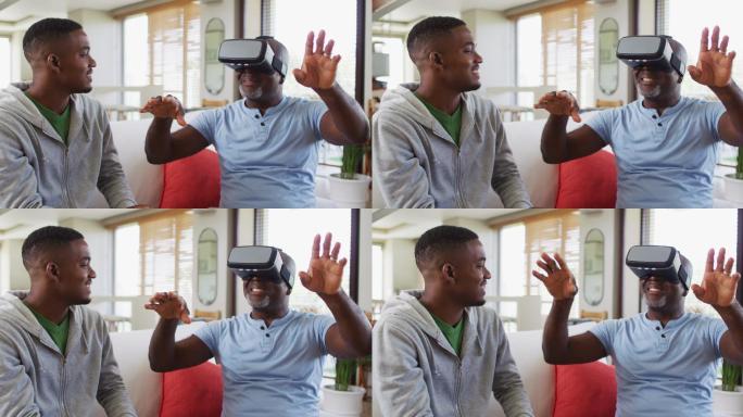一位非裔美国老人坐在沙发上，戴着虚拟现实头盔，他十几岁的儿子面带微笑