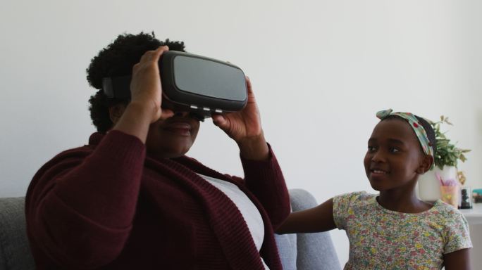 一位非裔美国祖母坐在家里的沙发上，使用虚拟现实头盔
