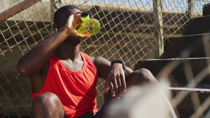 疲惫的非洲裔美国人坐着，喝着水瓶，在户外运动中休息