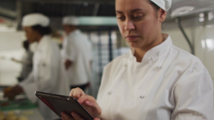 白人女厨师使用平板电脑在餐厅厨房与其他厨师的背景