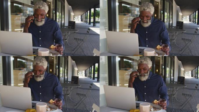 一位非裔美国老人坐在户外的咖啡馆里一边吃早餐一边用智能手机聊天