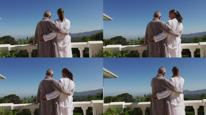 身穿浴袍的白人老年夫妇在自家阳台上拥抱对方的背影