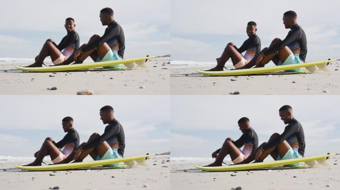 快乐的非洲裔美国青少年双胞胎兄弟坐在海滩上的冲浪板旁聊天