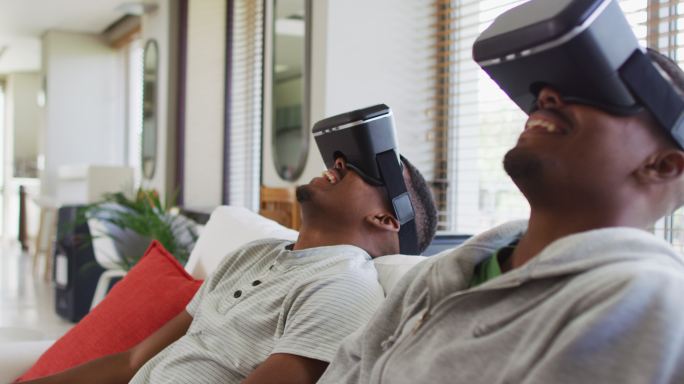 两个非洲裔美国青少年双胞胎兄弟坐在沙发上，戴着虚拟现实头盔微笑