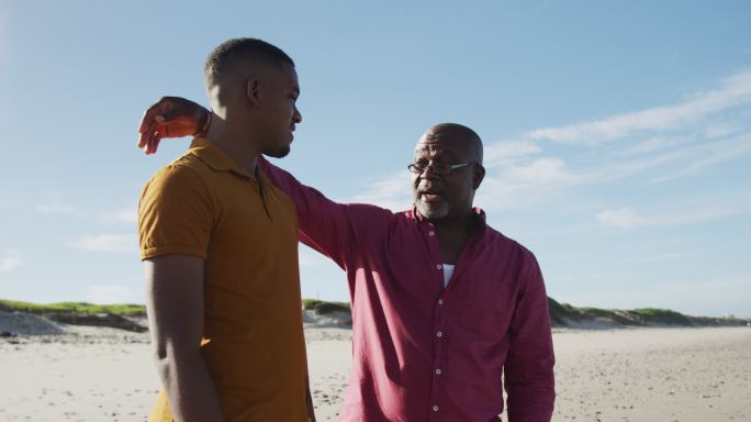 一位非裔美国父亲站在沙滩上，手搭在十几岁儿子的肩膀上说话