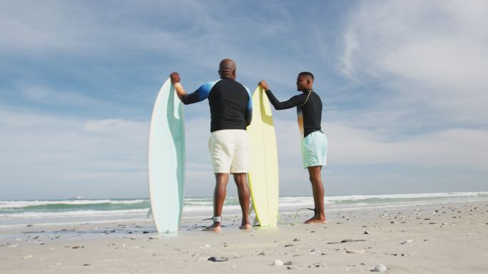 非裔美国父亲和十几岁的儿子站在海滩上，手里拿着冲浪板聊天