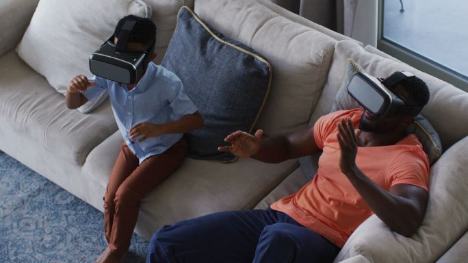 非裔美国人父亲和儿子一起在沙发上使用虚拟现实头盔