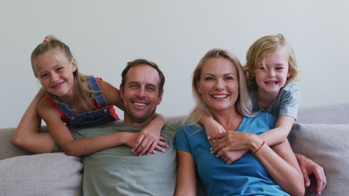 微笑的白人父母在沙发上与儿子和女儿拥抱他们的肖像