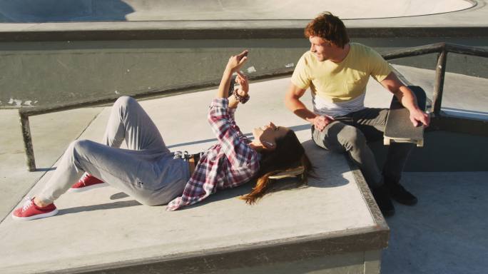 快乐的白人男女朋友在滑板场玩智能手机
