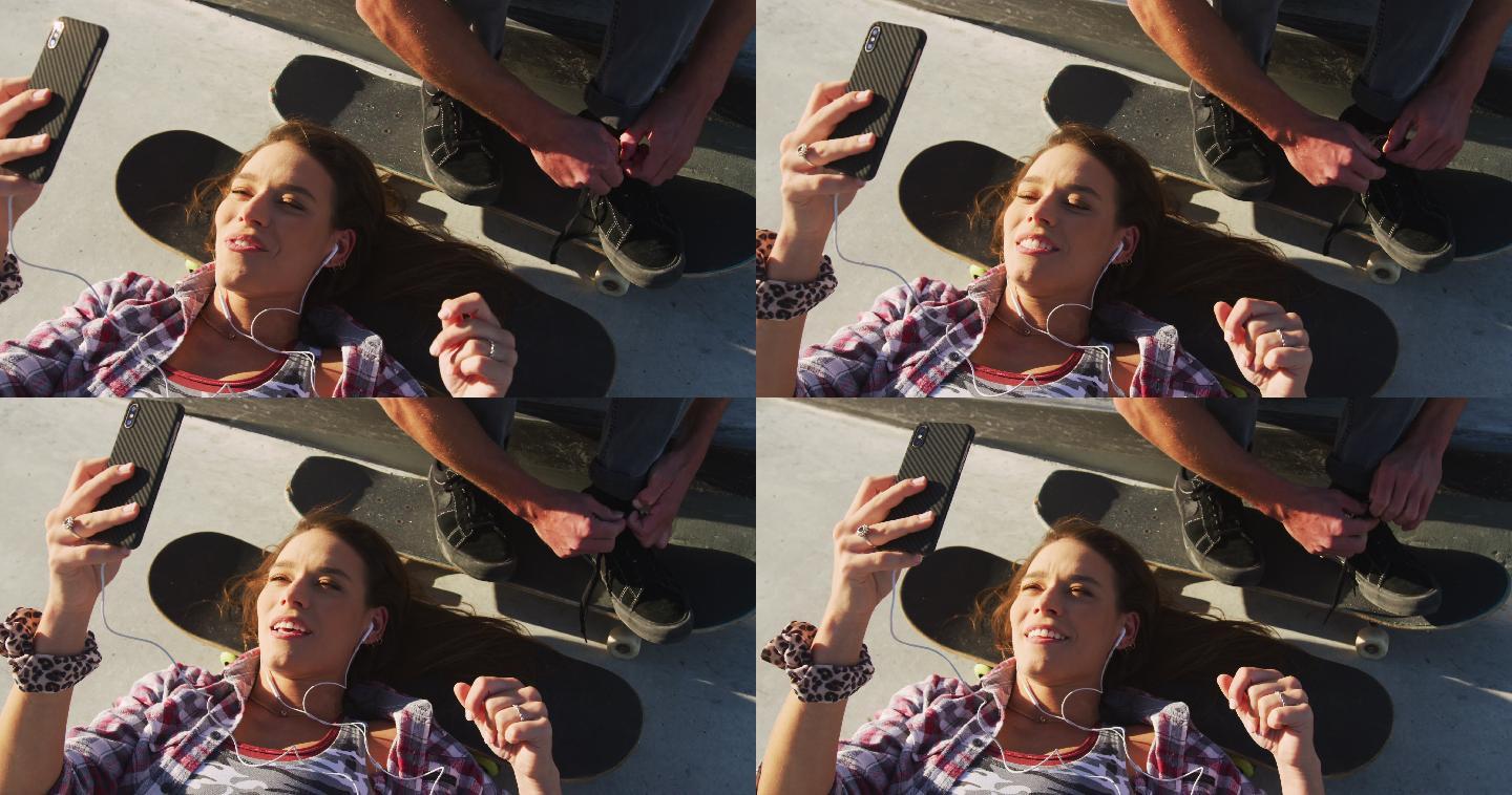 快乐的白人女人在玩智能手机，男人在滑板场系鞋带
