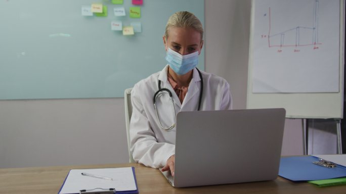 白种女医生戴着口罩，坐在会议室的桌子上用笔记本电脑