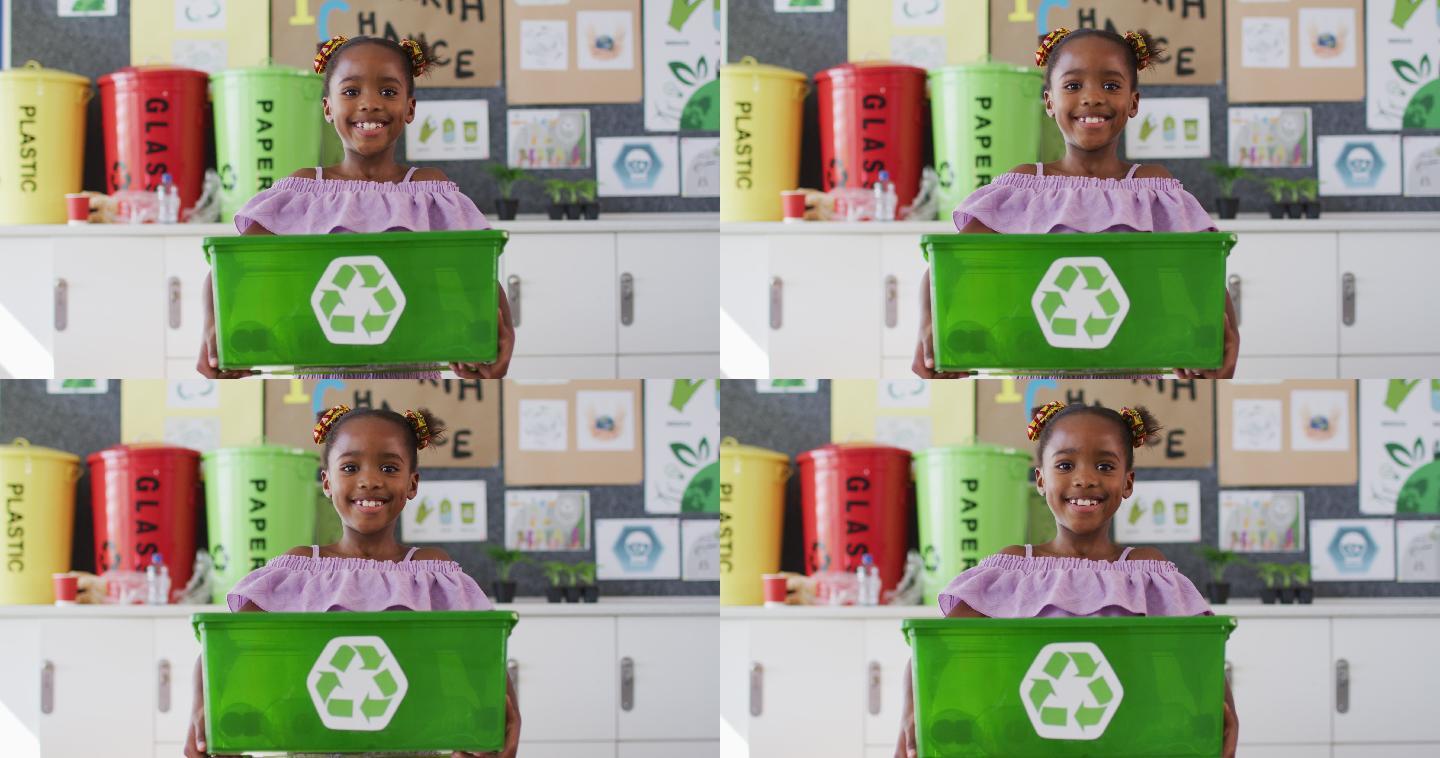 快乐的非裔美国女学生站在教室里，微笑着，拿着回收箱