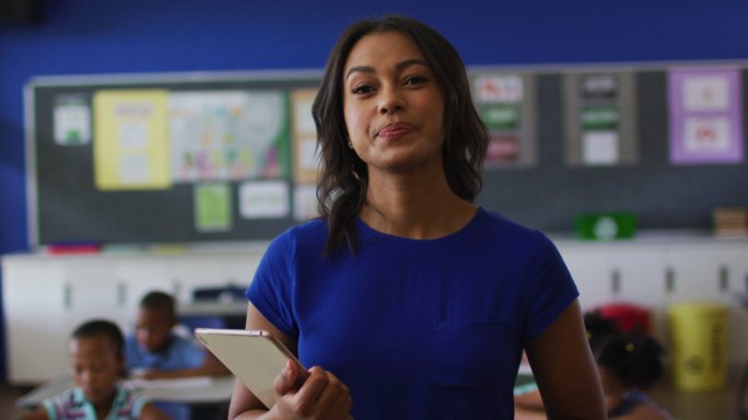 快乐的混血女教师的肖像站在教室里，背景是平板电脑的孩子们