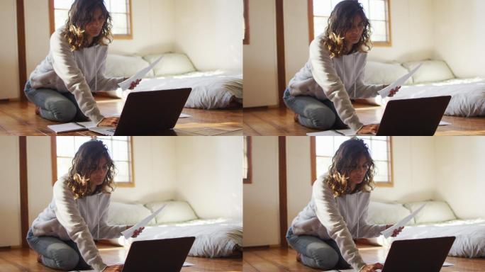 混血妇女在家工作，跪在地板上使用笔记本电脑，在小屋拿着文件