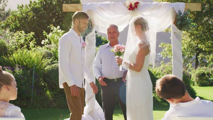 在婚礼仪式上，白人新娘和新郎站在室外的圣坛上，由婚礼司仪主持
