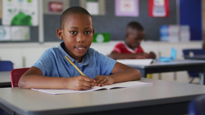 快乐的非裔美国学生的肖像坐在教室里，做笔记，看着相机