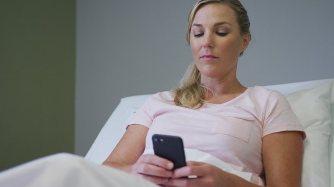 白人女性患者躺在医院病床上，使用智能手机
