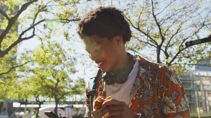 快乐的非裔美国人在城市，使用智能手机，戴着耳机，拿着糕点在街上