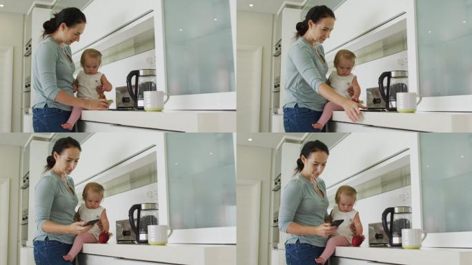 在厨房里抱着孩子用智能手机的白人母亲